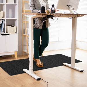 A person stands at an adjustable standing desk, working on a computer. The desk has various items on it, and the person is comfortably positioned on a TuffComfort Standing Desk Mat. A shelf with storage baskets is in the background.