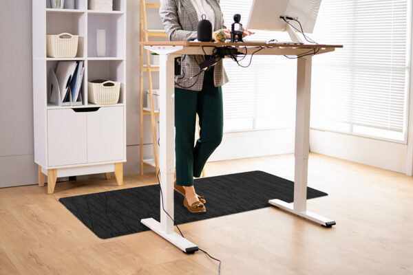 A person stands at an adjustable standing desk, working on a computer. The desk has various items on it, and the person is comfortably positioned on a TuffComfort Standing Desk Mat. A shelf with storage baskets is in the background.