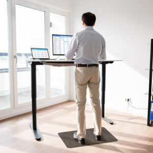 A person stands on a TuffComfort Standing Desk Mat at a height-adjustable desk in a bright room with large windows, working on a computer with multiple screens and surrounded by organized shelves with binders.