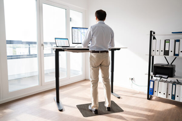 A person stands on a TuffComfort Standing Desk Mat at a height-adjustable desk in a bright room with large windows, working on a computer with multiple screens and surrounded by organized shelves with binders.