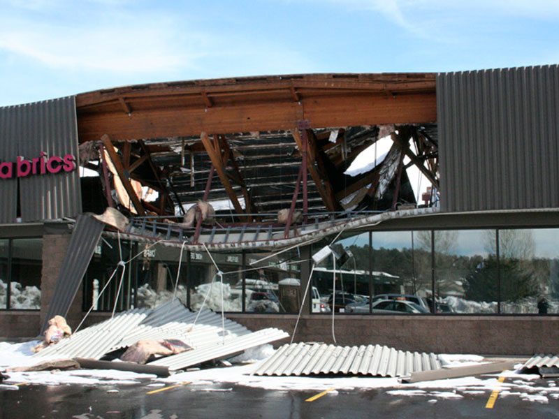 The collapsed roof of a commercial building, lacking a Roof Mat System, left debris scattered across the snow-covered ground. Large wooden beams lay exposed, with part of a store sign still visible—a testament to the unforeseen roof collapse.