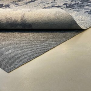 A grey fabric rug is partially rolled up, revealing its Teebaud underside on a smooth, beige floor.