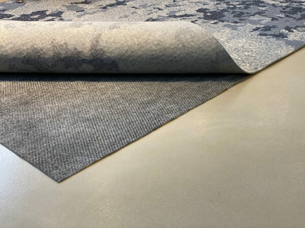 A grey fabric rug is partially rolled up, revealing its Teebaud underside on a smooth, beige floor.